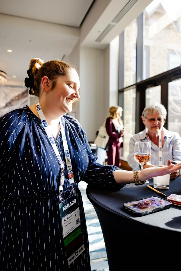 A conference attendee talks with a colleague at a daylit table.