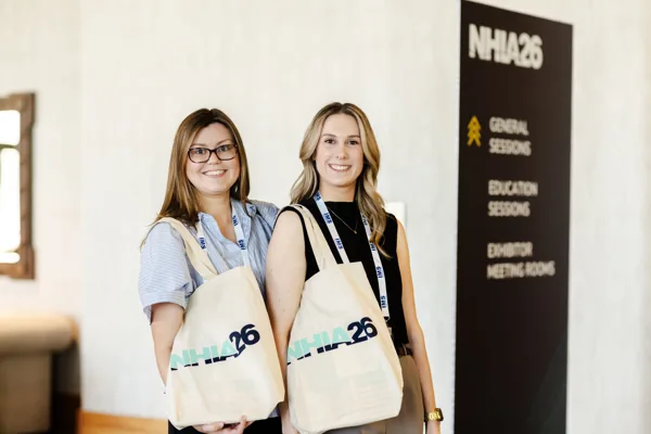Two attendees hold NHIA26 canvas tote bags beside a conference wayfinding sign.