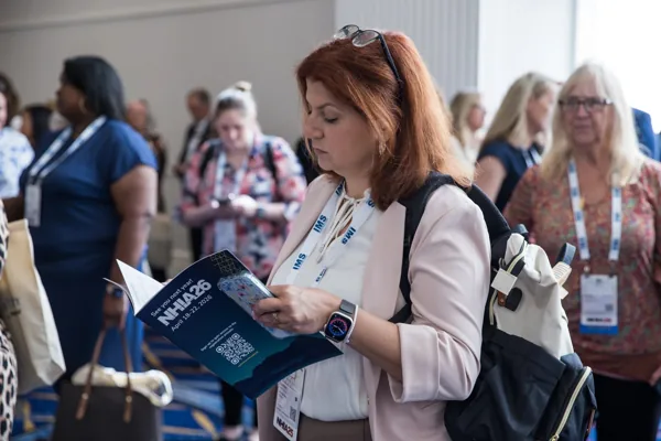 An attendee pauses in a busy hallway to read the printed Onsite Program, its back cover announcing NHIA26.