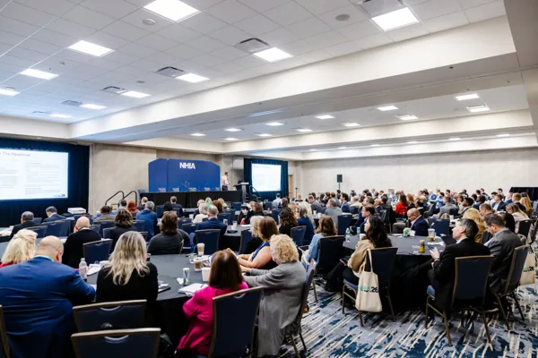 Attendees seated at round tables watch a presentation in a preconference session room.